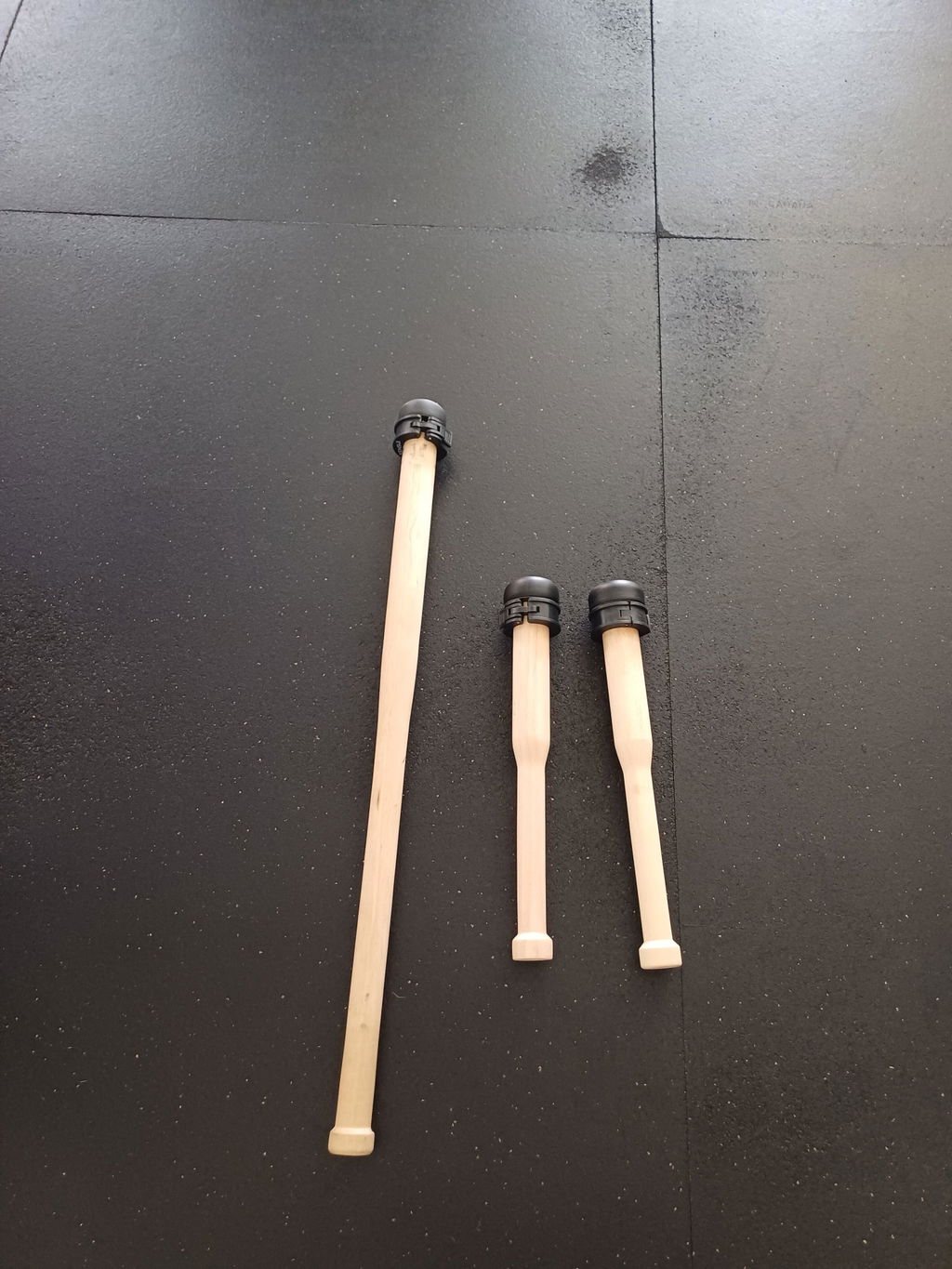 An owner's pale wooden mace lying on a black rubber gym floor beside two shorter wooden clubs, each with a black collar at its end
