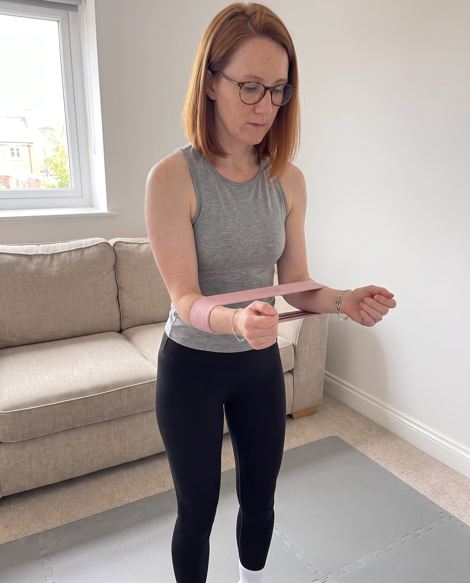 A woman in her living room with a pink fabric band around both wrists, elbows tucked, turning her forearms outward