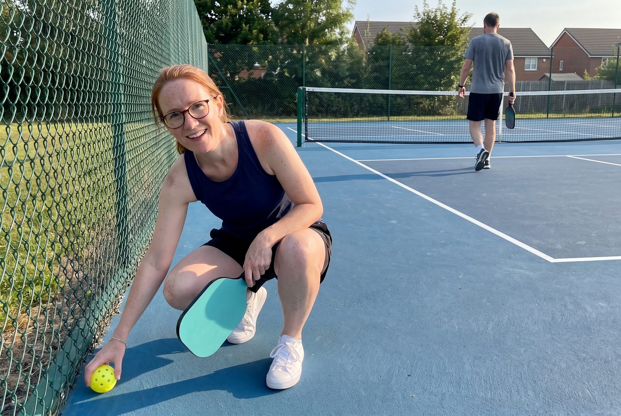 A woman with auburn hair and glasses crouches in the corner of a pickleball court to pick up a ball and smiles at the camera while a man walks back to his baseline