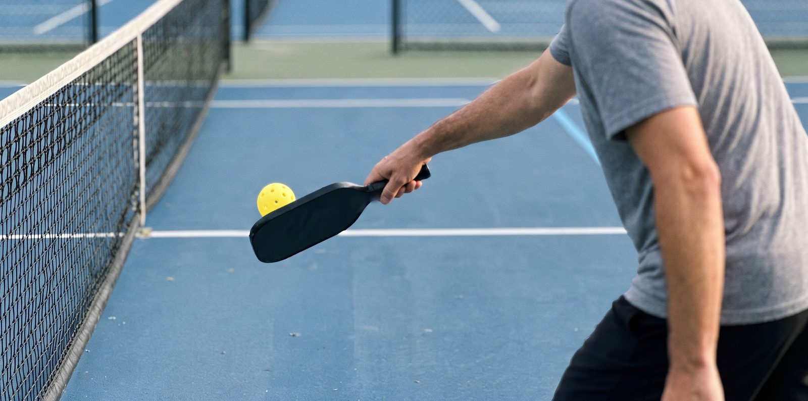 A man's arm, seen from behind, rolls a low backhand dink over the net with a loose grip on a black paddle