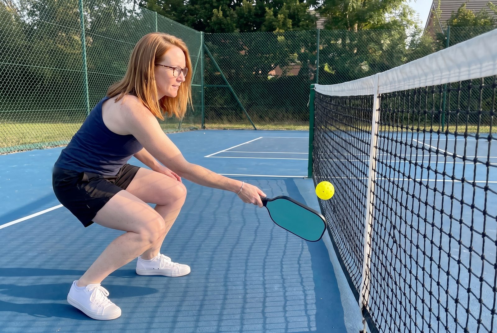 A woman crouches low at the net and hits a soft backhand dink that skims just over the tape