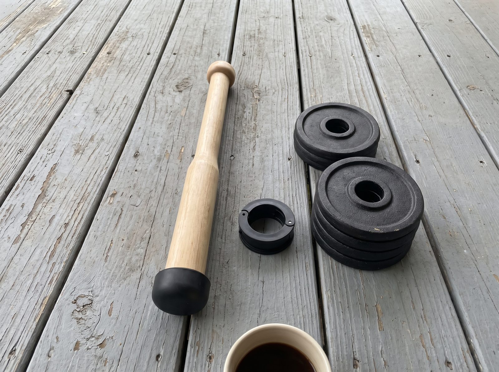 An empty wooden club, an open black collar and two small stacks of black weight plates on porch boards beside a coffee mug