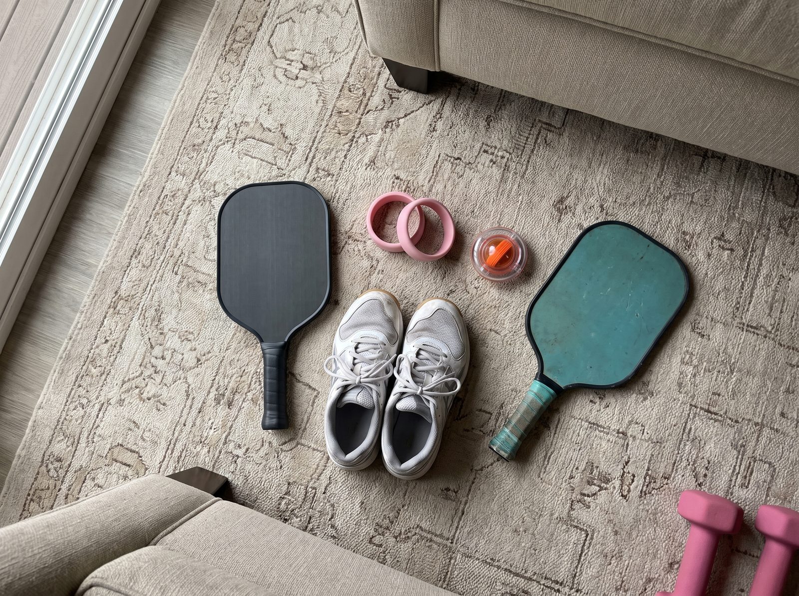 A charcoal paddle, court shoes, pink wrist weights, a gyro wrist ball and an old teal paddle laid out on a living room rug next to pink dumbbells