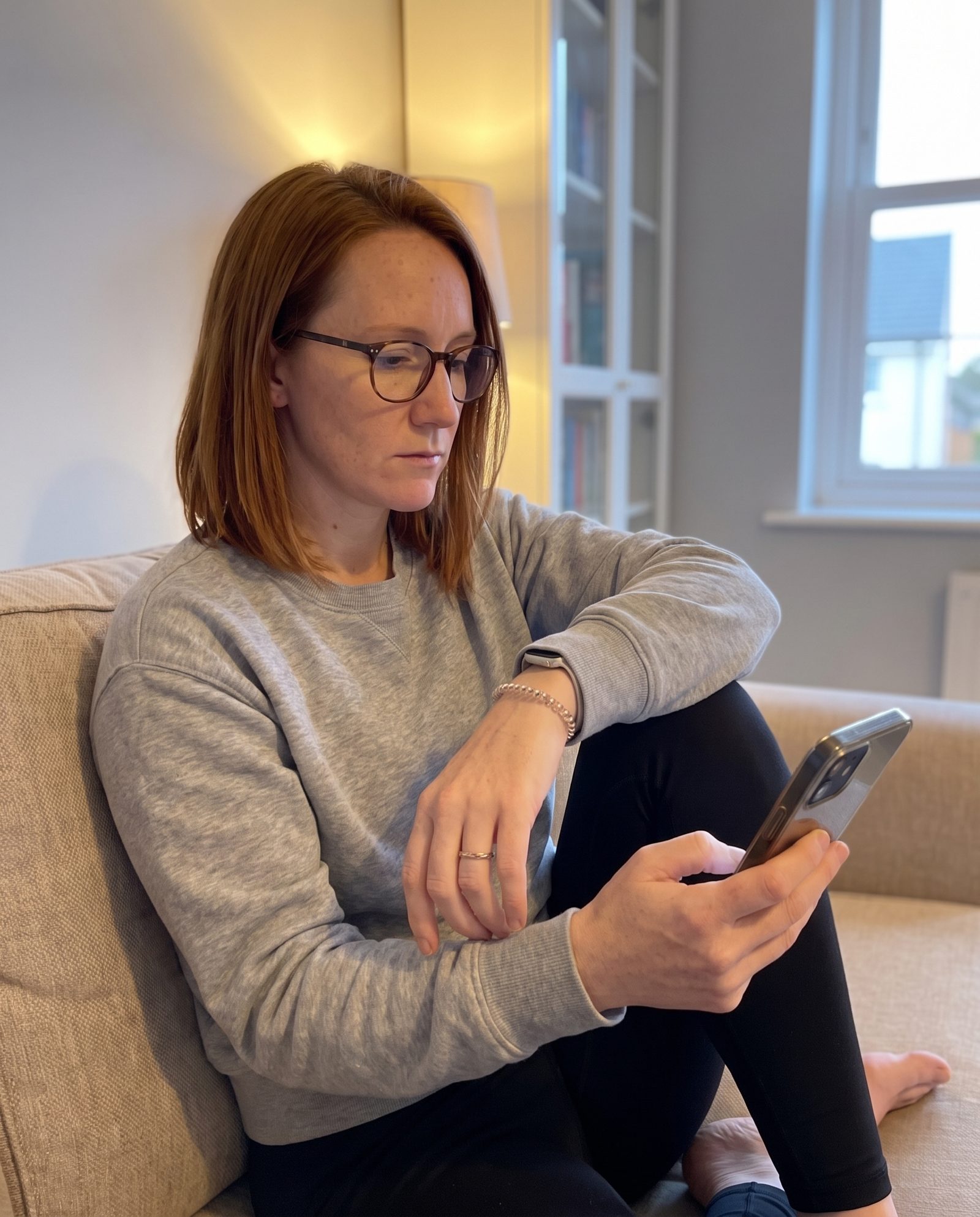 A woman with auburn hair and glasses sits on a sofa reading her phone, her other wrist resting on her knee