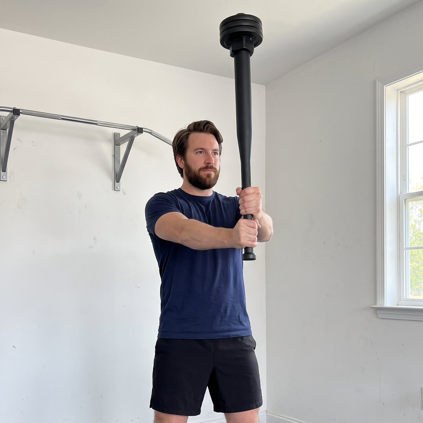 A man in a navy t-shirt in a white garage holding a long black steel plate-loaded club upright with both hands, a stack of plates locked on the top end above his head