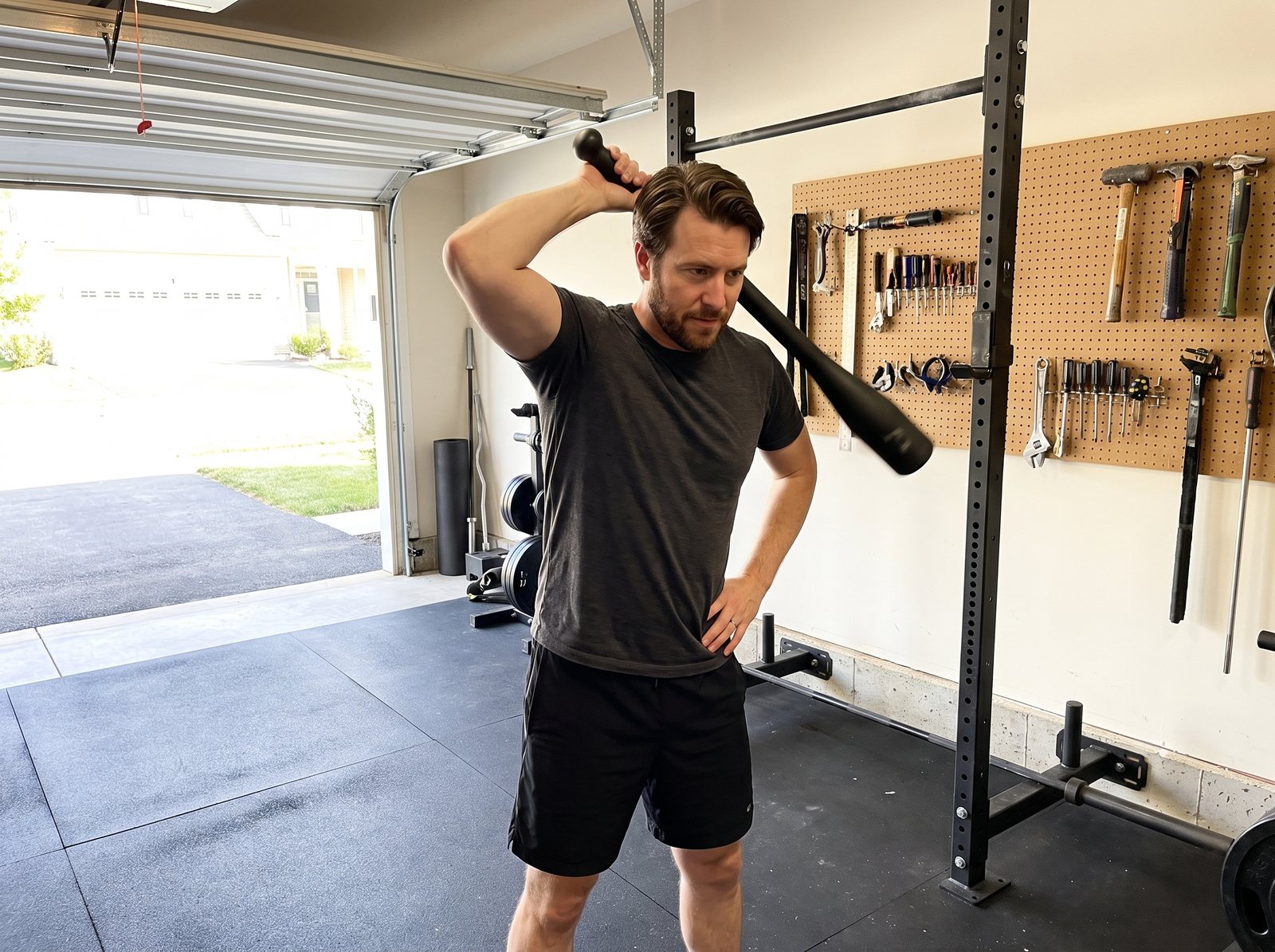 A man in a garage gym halfway through a club mill, one hand holding a black steel club beside his head with the heavy end hanging behind his shoulders