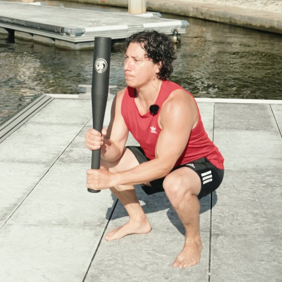 A man in a deep squat on a dock holding a black steel club upright in front of his chest