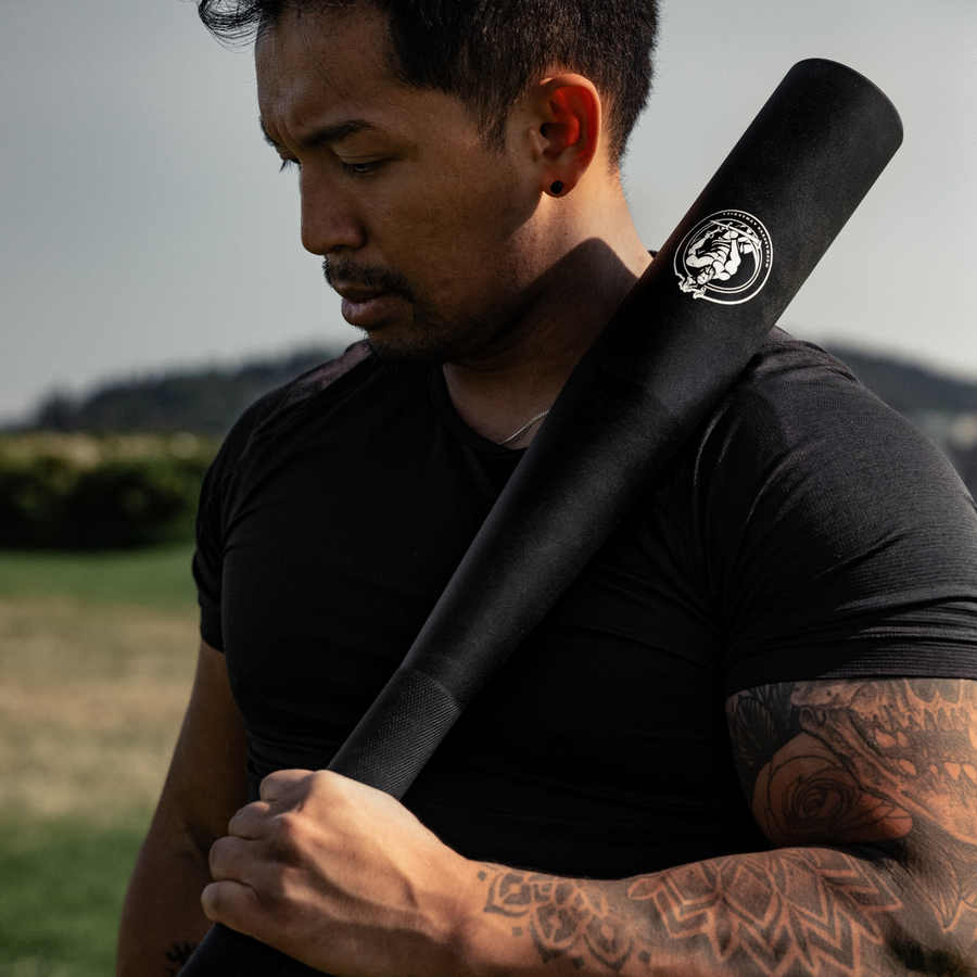 A man in a black t-shirt outdoors resting a black fixed steel club on his shoulder