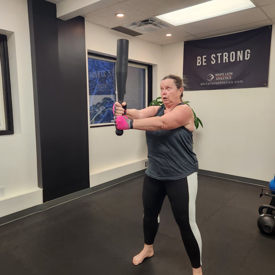A woman in a small gym holding a black fixed steel club upright in front of her with both hands