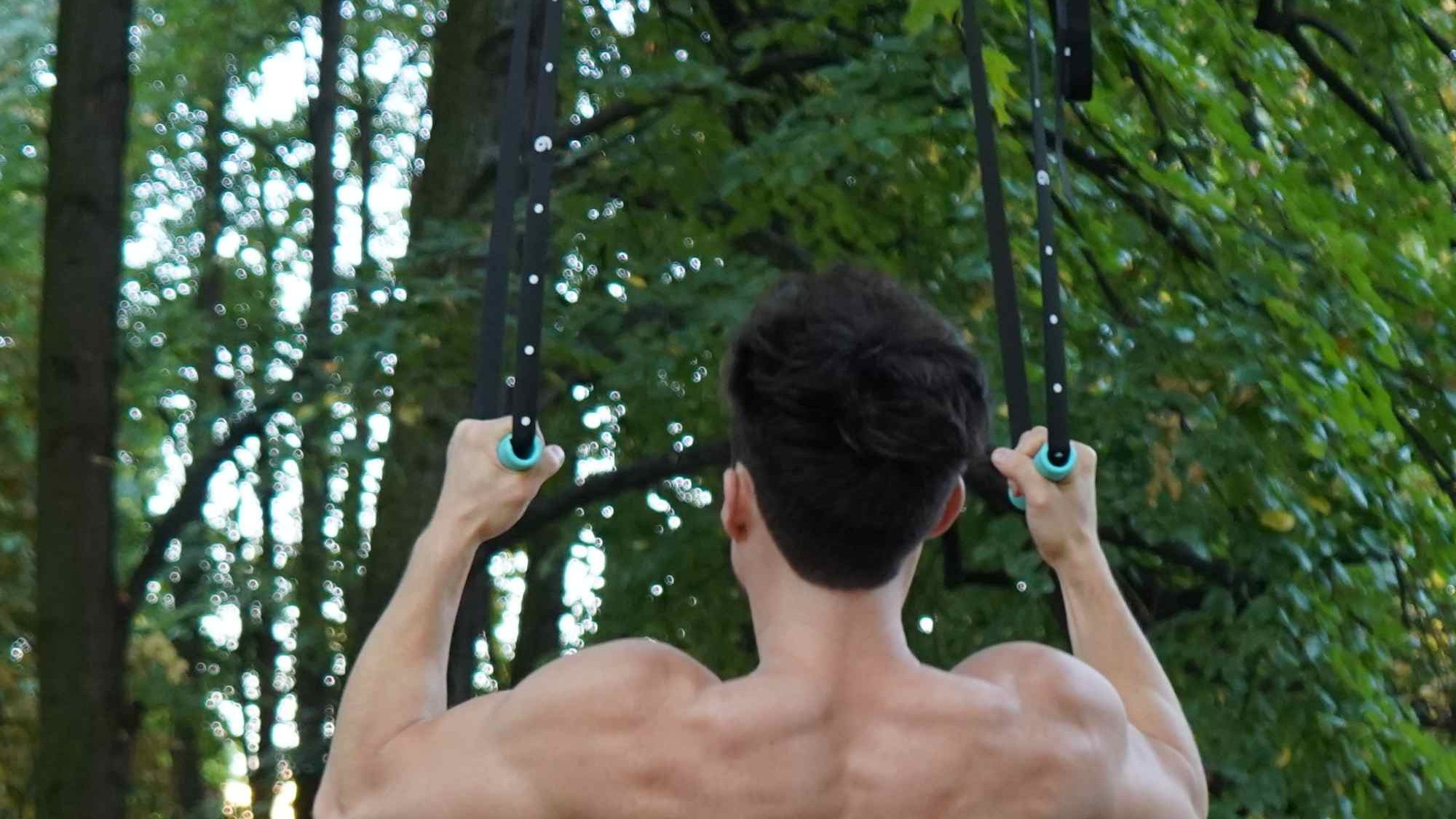 A man seen from behind pulling up on a pair of Kensui Nanobars V2 suspension straps hung from a tree