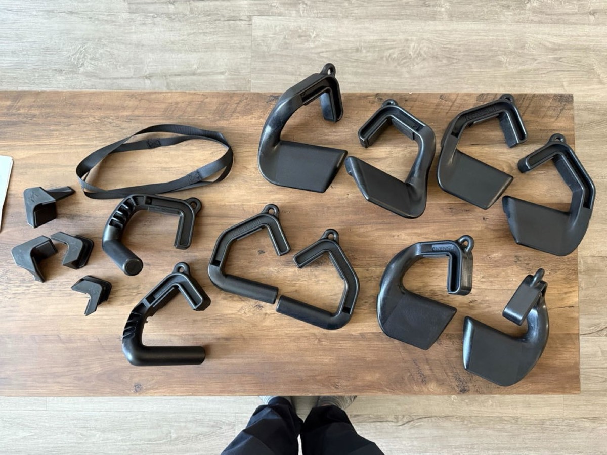 Overhead owner photograph of a wooden table covered with more than a dozen Kensui grip handles and hook attachments of different shapes, plus a nylon strap, shot looking down past a pair of feet.