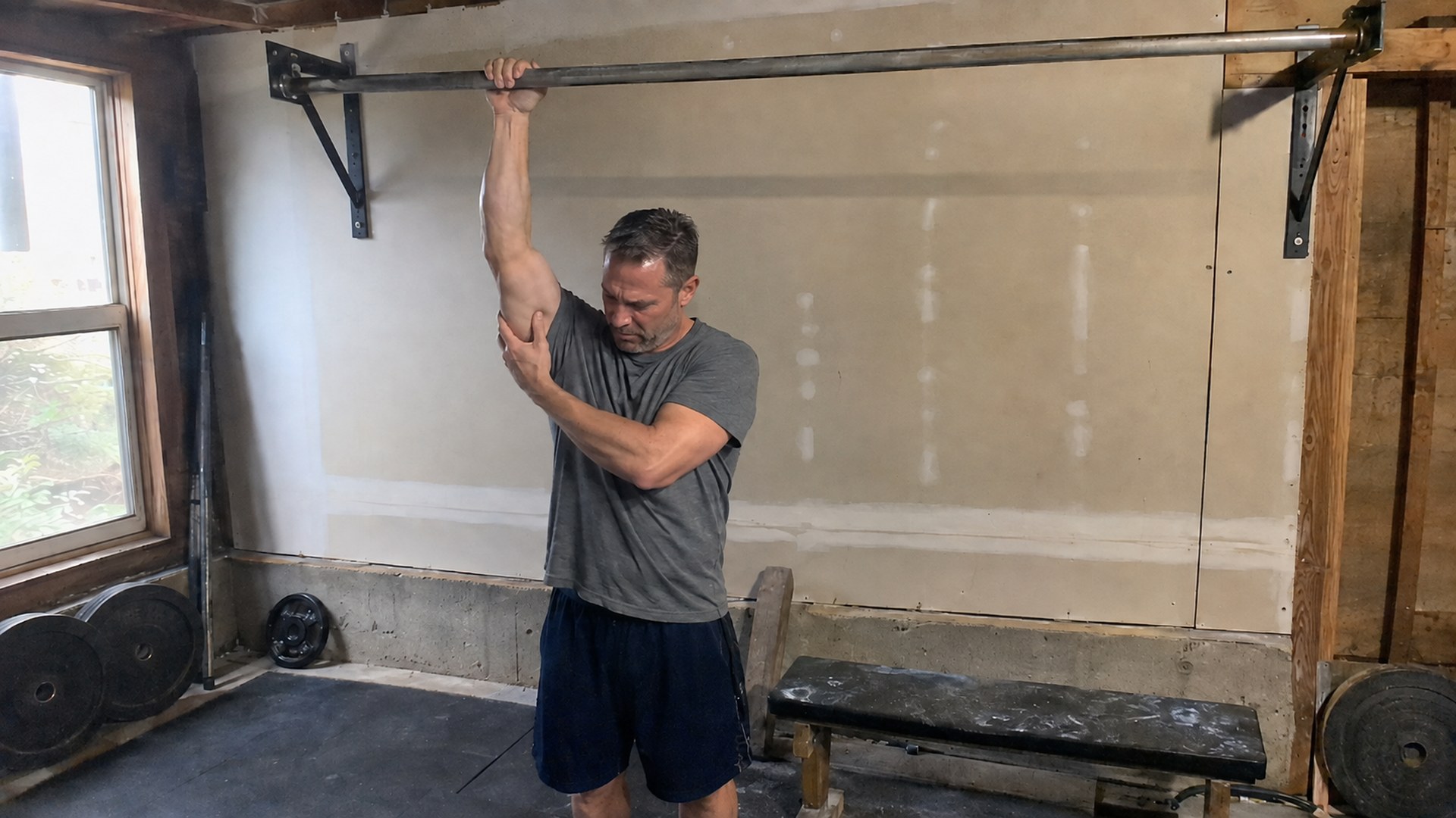 A lifter standing under a home pull-up bar, one hand still on the bar, squeezing his own forearm below the elbow