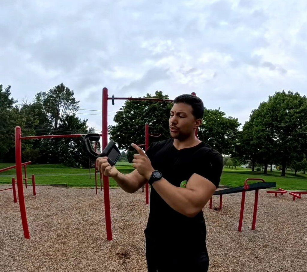 Owner photograph at an outdoor street-workout park: a man in a black t-shirt holds a pair of Kensui Swissies handles up in front of a red steel calisthenics rig, about to hang them on the bar behind him.