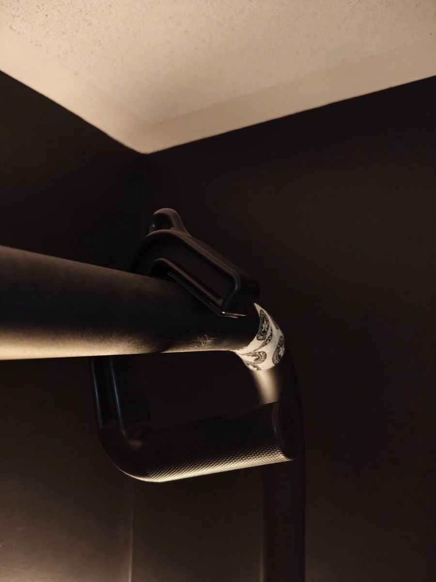 Owner photograph in a dimly lit room: a single Swissies V2 handle hooked over a thick black pull-up bar, showing the two contact points of the hook wedged against the bar.