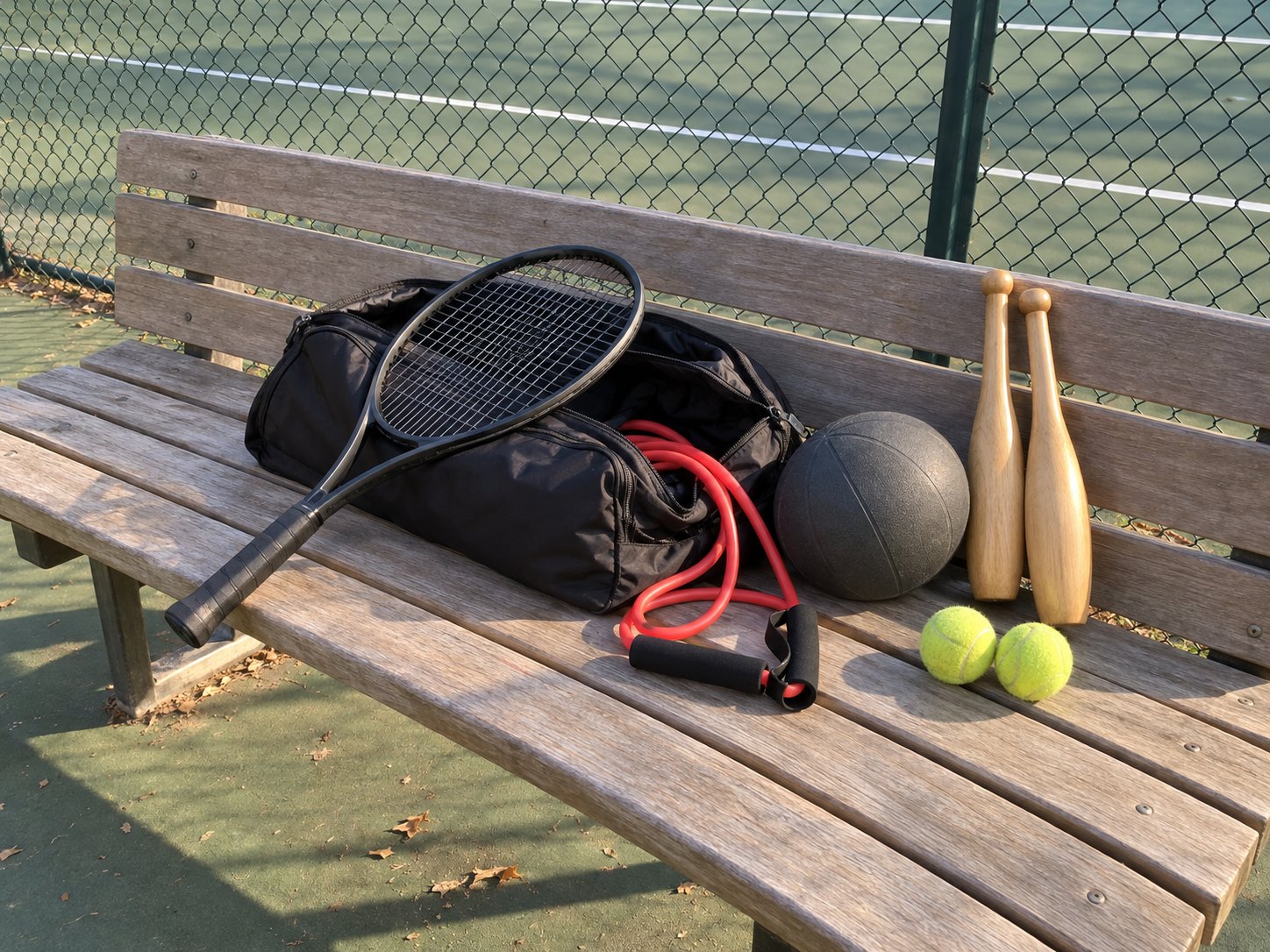 An open tennis bag on a court bench with a coiled red resistance cord, a grey medicine ball and two light wooden clubs spilling out beside a racquet
