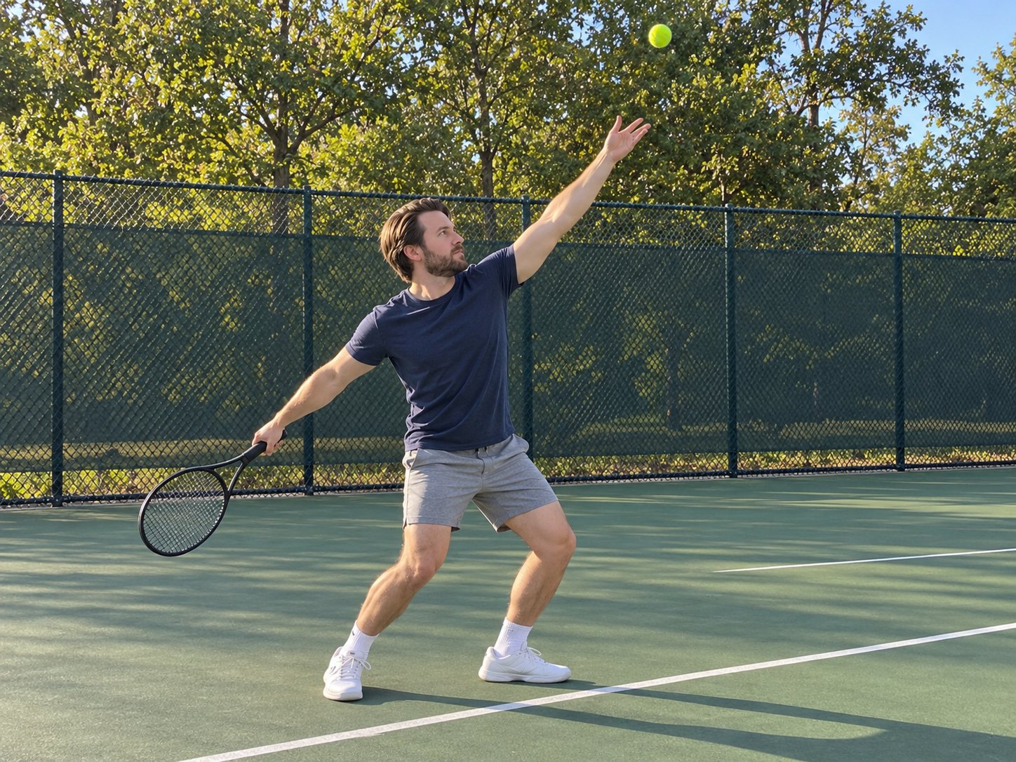 A man in a navy t-shirt on a public hard court starting a serve, the ball tossed high above his outstretched arm and the racquet low behind him