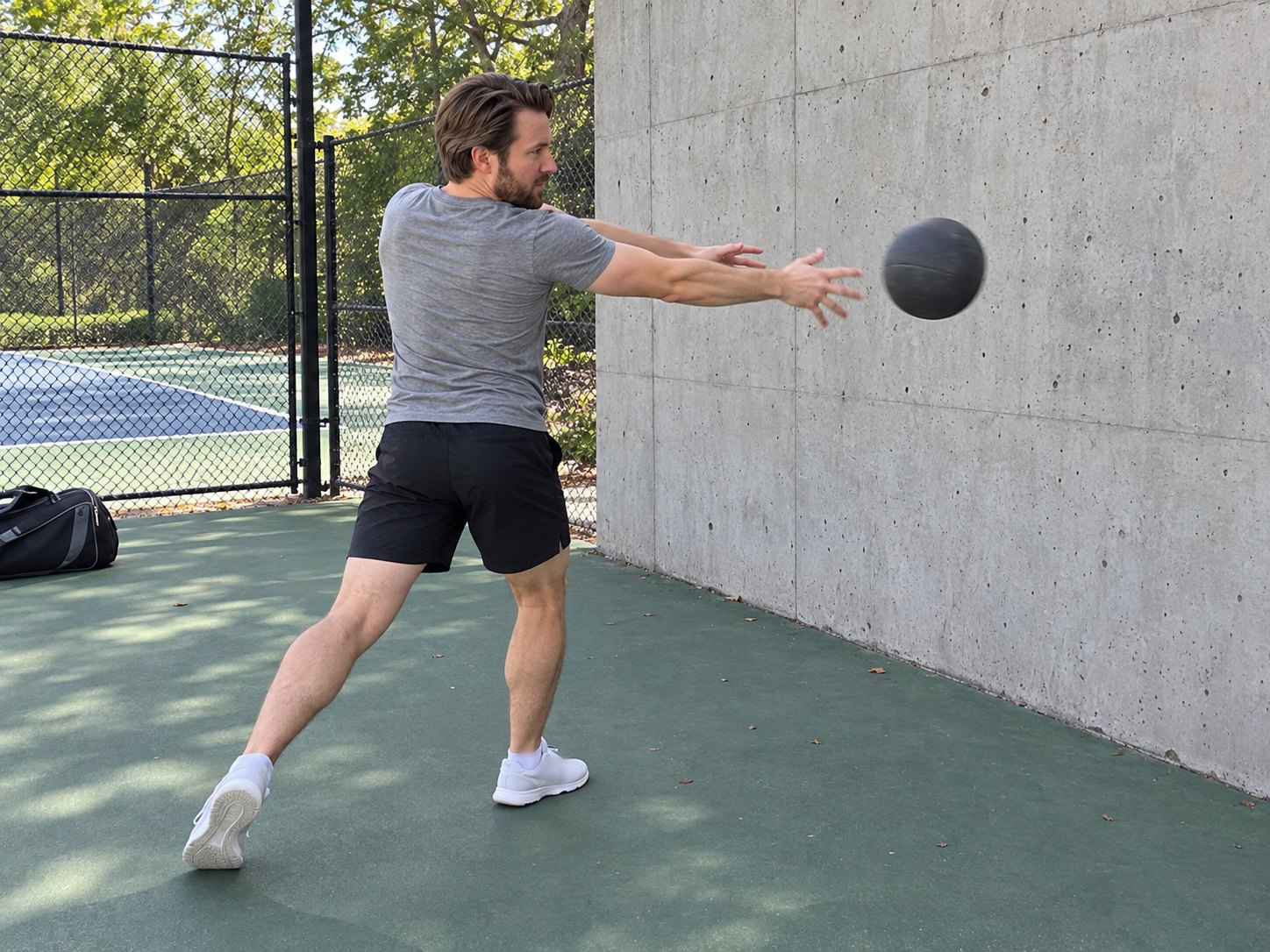 A man beside a tennis court twisting hard to throw a grey medicine ball at a concrete practice wall, his back heel lifted as the ball leaves his hands