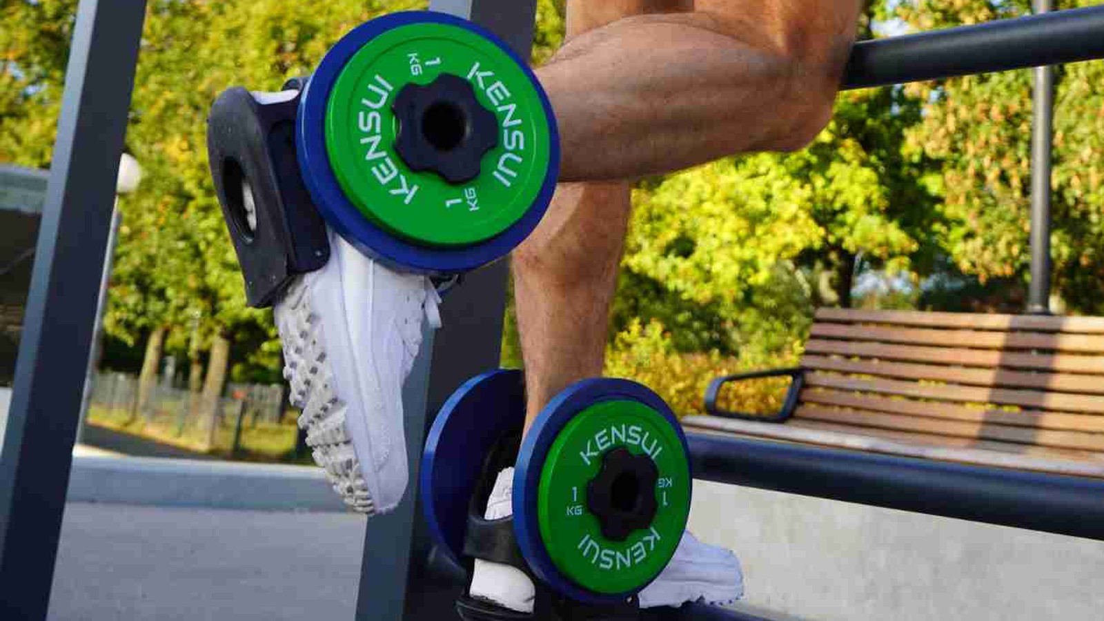 A man doing a lower leg exercise on an outdoor bar with green weight plates strapped to both feet in plate-loaded boots