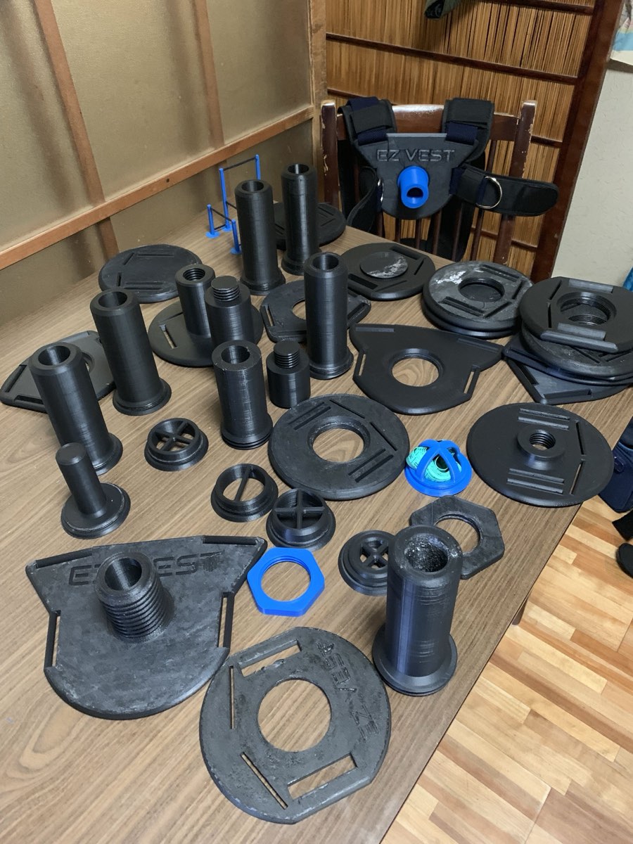 A dining table covered in black 3D printed loading posts, collars and plate adapters, with an early vest propped on the chair behind it