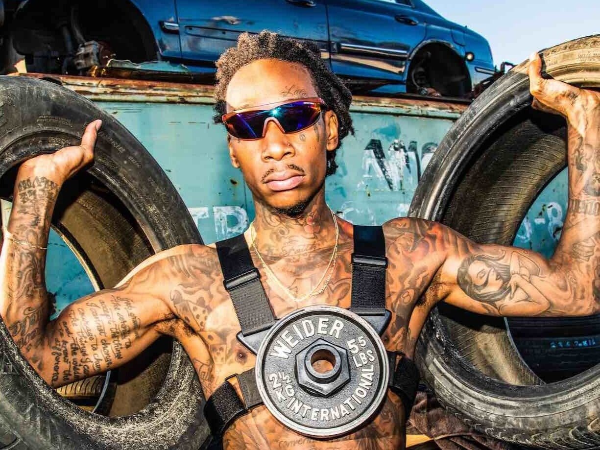 Wiz Khalifa in a scrapyard holding a tire in each hand, wearing a black two-strap chest rig with one 5.5 pound Weider plate